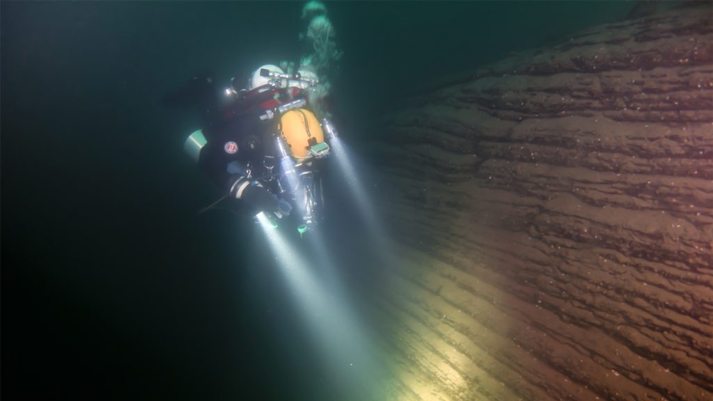 Subacqueo in configurazione tecnica illumina una parete stratificata nel Lago di Como durante un’immersione profonda