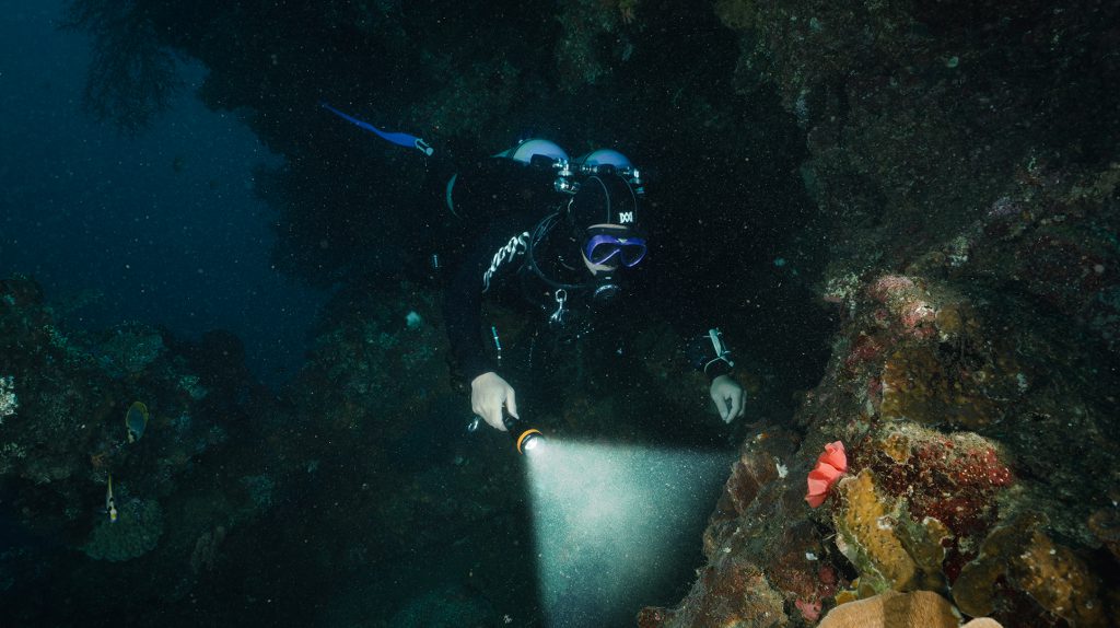 Subacqueo con attrezzatura tecnica esplora un relitto con torcia durante un'immersione in acque profonde.