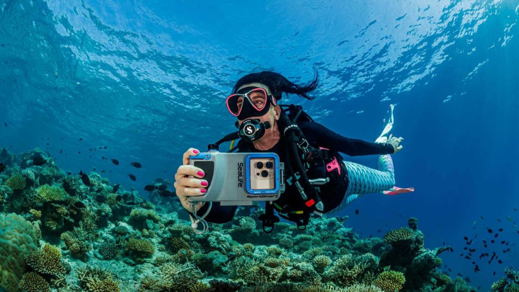 Subacquea in immersione con custodia Sealife SportDiver S per smartphone, mentre fotografa una barriera corallina tropicale.
